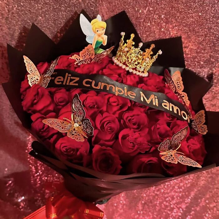 A large bouquet of deep red roses in black paper, topped with a gold pearl crown and a Tinker Bell figure, rose-gold filigree butterflies pinned across the blooms and a black sash lettered in gold reading Feliz cumple Mi amor.