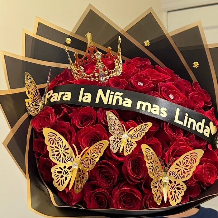 A bouquet of red roses in a star of black and gold paper points, with a gold crown set into the blooms, large gold filigree butterflies and a black sash lettered Para la Ni&ntilde;a mas Linda.