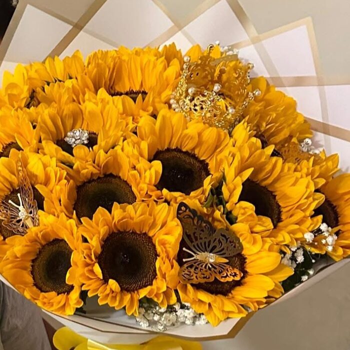 A dense round bouquet of about twenty sunflowers in white and kraft wrapping paper, with a gold pearl crown set among the blooms, two filigree butterflies and sprigs of white gypsophila.